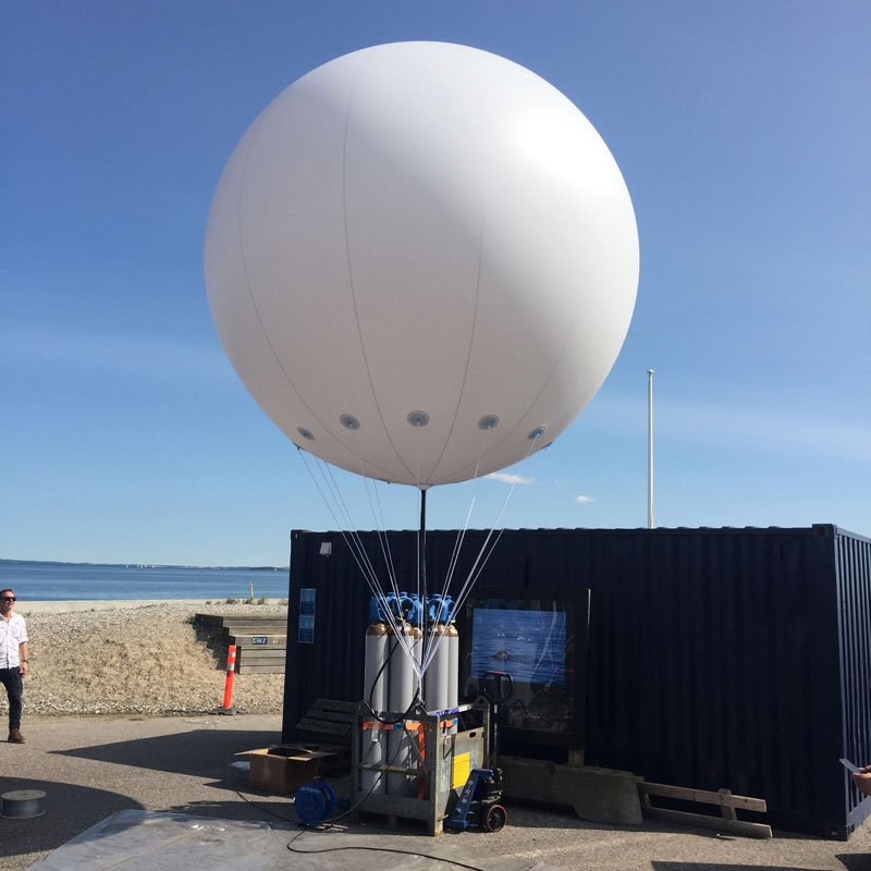 Ballon blanc de grand diamètre relié à plusieurs câbles devant un conteneur en extérieur
